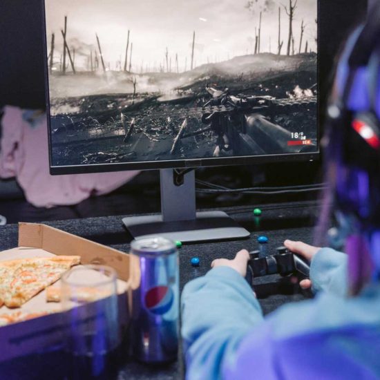 Teen playing video game with headset and drinks, indulging in pizza during gaming session.